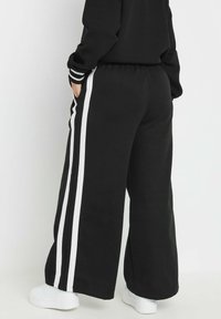 Pantalons larges noirs en matière douce, avec des rayures blanches sur les côtés, une taille élastique et des poches latérales. Portés avec des baskets blanches.