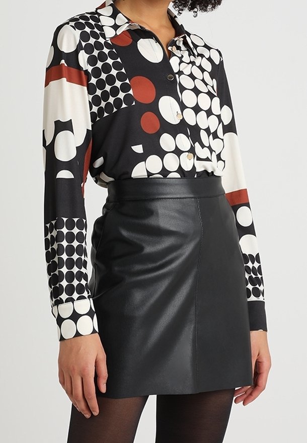 Žena v černé kožené mini sukni s černými punčochami a košilí s dlouhým rukávem, která má černé, bílé a červené kruhové vzory.