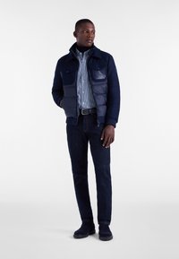 Granatowa kurtka puchowa z kołnierzykiem i dwiema kieszeniami na piersi, założona na jasnoniebieską koszulę w kratę, w komplecie z ciemnymi dżinsami i granatowymi butami.