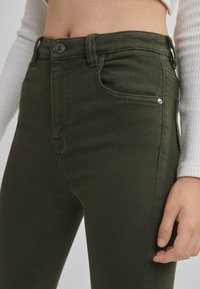 Gros plan sur une personne portant un jean taille haute vert olive et un haut blanc moulant à manches longues, montrant le torse et les hanches.