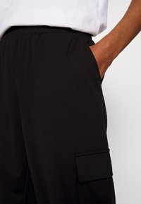 Pantalon noir en tissu doux, avec une taille élastique, des poches latérales et une poche plaquée sur la cuisse gauche.