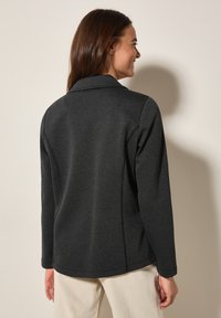 Veste grise texturée avec une coupe cintrée et des manches longues, dotée d'un col à cran et de détails de couture, portée sur un pantalon de couleur claire.