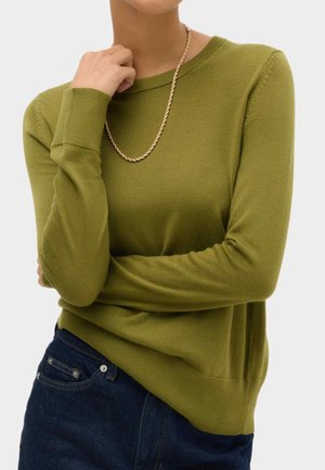 Frau trägt olivgrünen Langarmpullover, dunkelblaue Jeans und eine gedrehte goldene Kette, mit einer Hand nahe am Hals.
