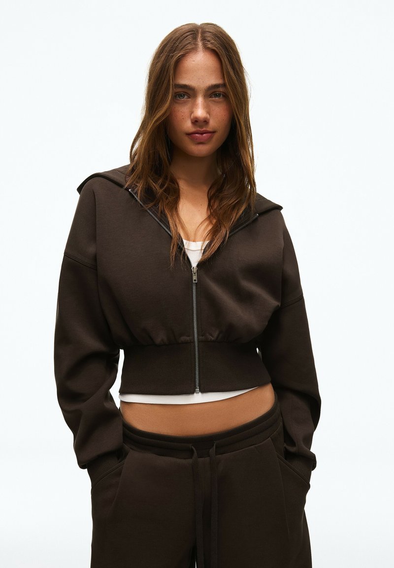 Jeune femme aux longs cheveux bruns portant un sweat à capuche court zippé marron foncé et un pantalon de jogging assorti, debout, les mains dans les poches.