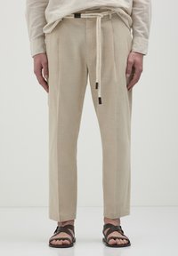 Beige, strukturierte Hose mit geradem Schnitt, ausgestattet mit einem Seilgürtel und braunen Akzenten. Kombiniert mit dunklen Sandalen vor einem neutralen Hintergrund.