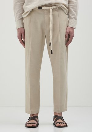 Beige, strukturierte Hose mit geradem Schnitt, ausgestattet mit einem Seilgürtel und braunen Akzenten. Kombiniert mit dunklen Sandalen vor einem neutralen Hintergrund.