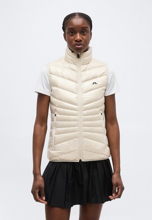 Μαύρη γυναίκα που φοράει μία εκρού γιλέκο με ραφές χωρίς μανίκια πάνω από μία λευκή κοντομάνικη μπλούζα και μια μαύρη πλισέ φούστα, στέκεται μπροστά από ένα απλό φόντο.