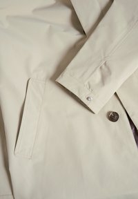 Bežna dežna plašč s gladko teksturo. Ima gumbasto zapiranje, stranske žepe in en sam gumb na manšeti. Minimalističen dizajn.