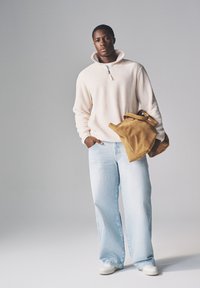 Beżowy polarowy sweter na pół z zamkiem, jasnoniebieskie luźne dżinsy, białe trampki i beżowa zamszowa torba typu tote, na tle szarego tła.