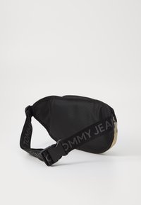 Sac banane noir en nylon avec un fond beige. Dispose d'une large sangle avec "TOMMY JEANS" imprimé en gris. Texture lisse et forme arrondie.