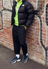 Svart puffjacka, neongrön hoodie, svarta byxor och marinblå sneakers med gröna accenter, står mot en tegelvägg.