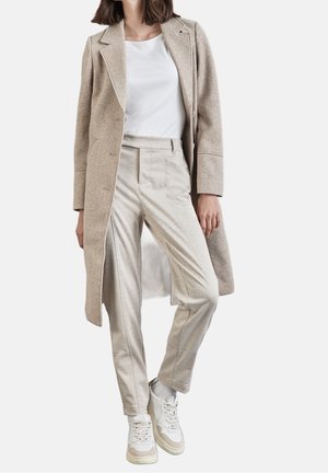 Mujer con abrigo largo beige, camiseta blanca, pantalones beige a medida y zapatillas blancas sobre un fondo blanco liso.