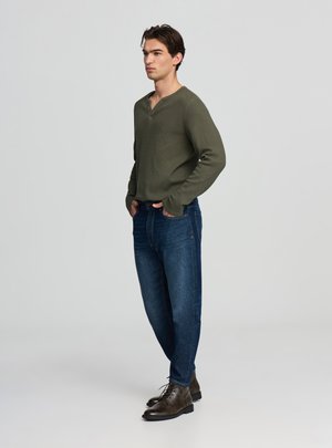 Maglietta henley a maniche lunghe verde oliva con una finitura testurizzata, abbinata a jeans di denim blu e stivaletti marrone scuro. Il modello è ritratto di profilo.