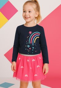 Dunkelblaues Langarmoberteil mit mehrfarbigem Glitzer-Regenbogen- und Sternendesign; kombiniert mit einem pinkfarbenen Tüllrock, der kleine Regenbogenmuster aufweist.