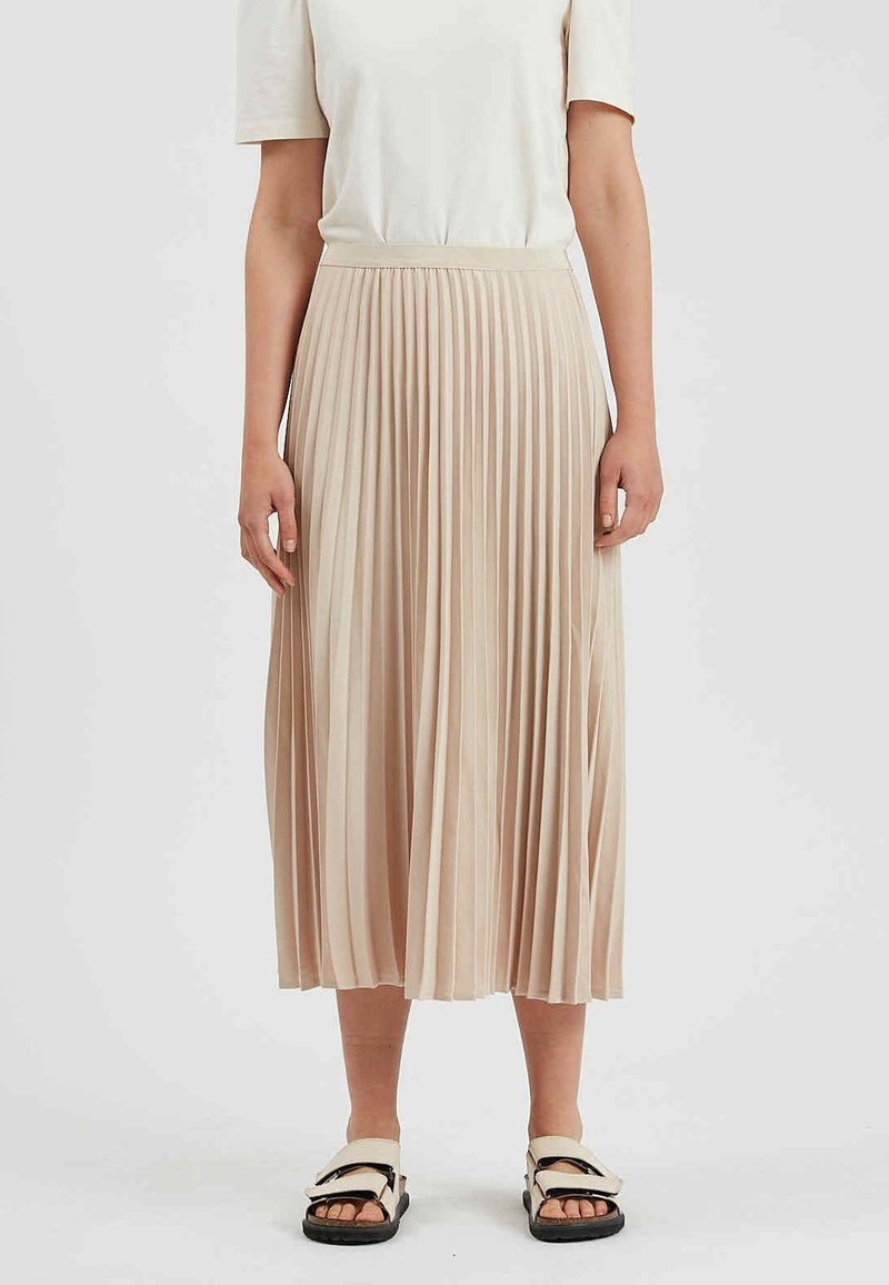 Femme portant un haut blanc à manches courtes, une jupe midi plissée beige et des sandales beiges avec semelles noires sur fond blanc.
