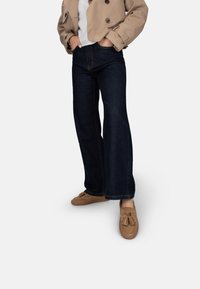 Jeans acampanados de color azul oscuro con costuras visibles y ajuste de cintura media, combinados con zapatos de estilo mocasín en color beige y una chaqueta corta de color beige.
