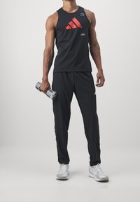 Černý bezrukávový sportovní top s červeným logem Adidas, v kombinaci s černými zúženými kalhotami a bílými teniskami. Osoba drží činky.