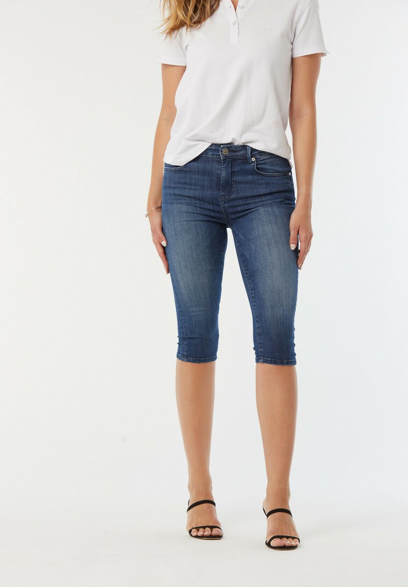 Jeans capri en denim bleu foncé avec un design ajusté, une coupe au genou et un léger délavage ; accompagnés d'un polo blanc et de sandales noires.