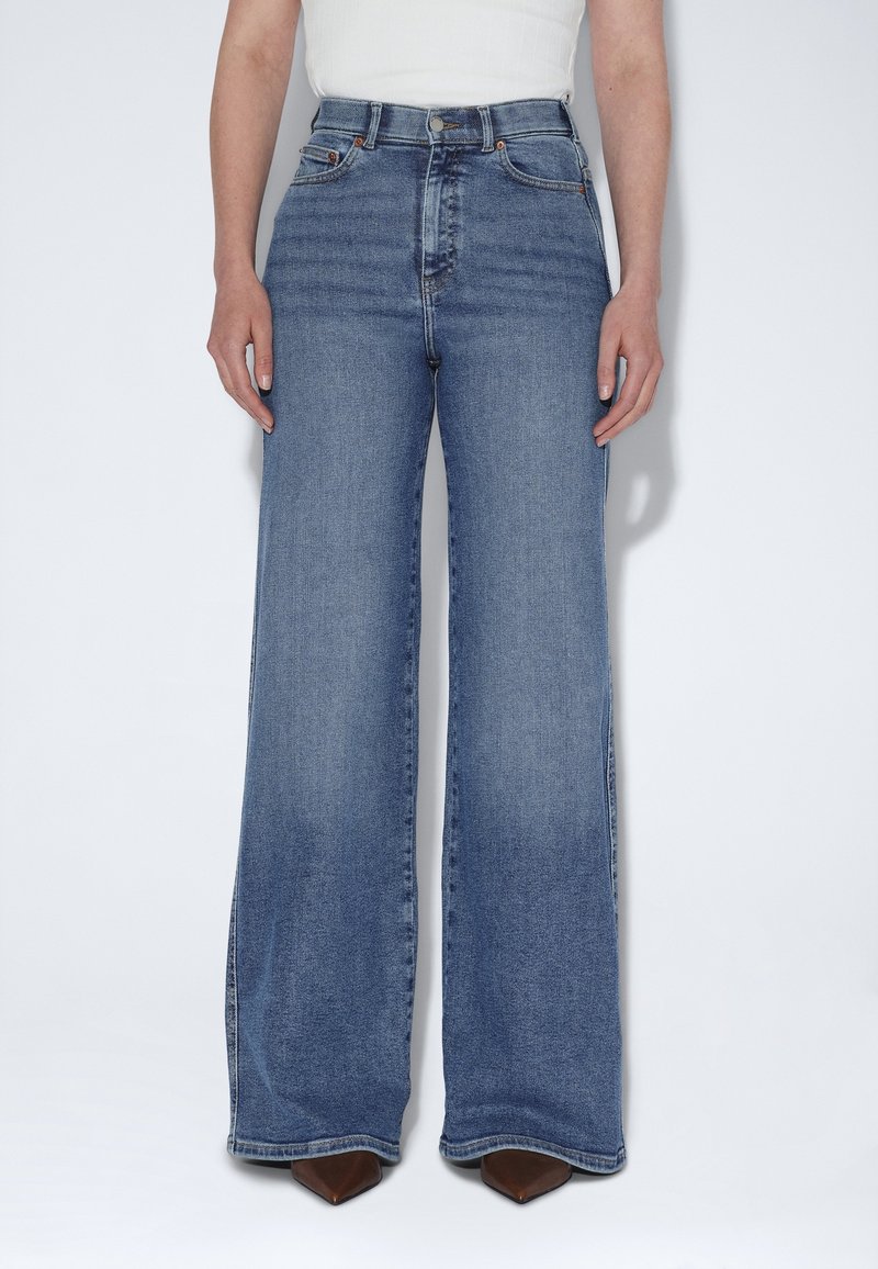 Personne portant un jean bleu taille haute à jambes larges et des chaussures marron à bouts pointus, debout devant un fond blanc.