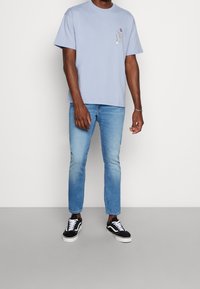 Man som bär en ljusblå grafisk t-shirt, ljusblå slim-fit jeans och svarta och vita sneakers, stående mot en enfärgad bakgrund.