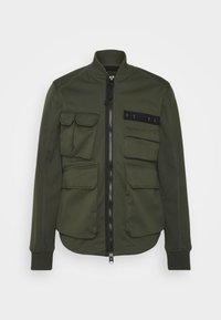 Oliwkowa kurtka bomberka z zamkiem z przodu, czterema przednimi kieszeniami, ściągaczami na rękawach oraz czarnym materiałowym akcentem z literami. Gładka faktura.