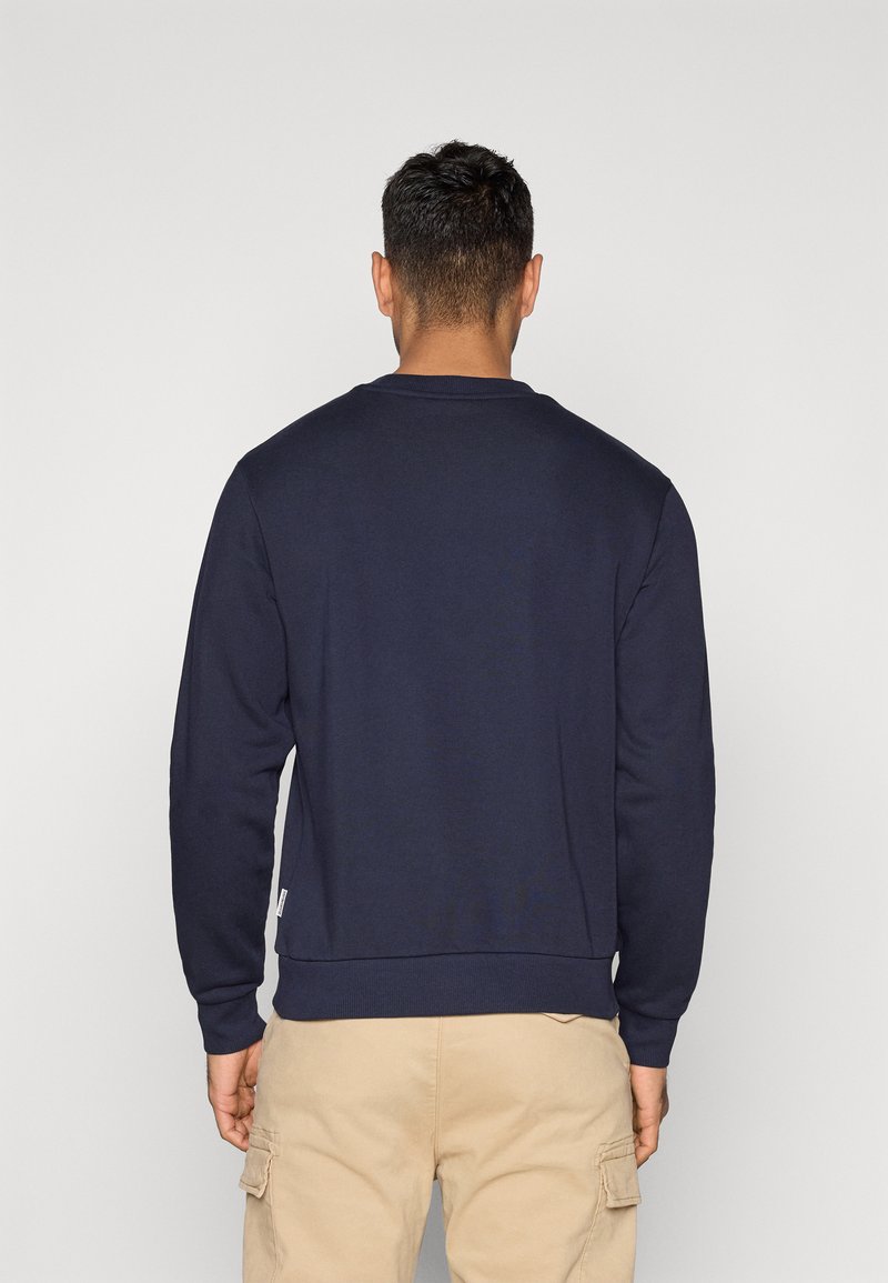 Un homme vu de dos portant un sweat-shirt à manches longues bleu marine et un pantalon cargo beige, sur un fond clair uni.