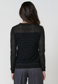 Una donna con capelli lunghi è vista di spalle, indossando un maglione nero a maglia testurizzata e pantaloni grigi, che mettono in evidenza un dettagliato motivo a maglia.