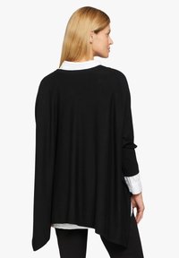 Garment in stile poncho di colore nero realizzato in tessuto morbido, caratterizzato da una vestibilità ampia e rilassata e una parte posteriore più lunga. Indossato sopra una camicia bianca con colletto.