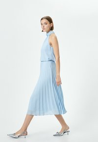 Vaaleansininen laskostettu mekko, jossa on korkea pääntie, yhdistettynä metallinhohtoisiin hopeisiin slingback-kengiin, joissa on terävät kärjet ja pienet korot.