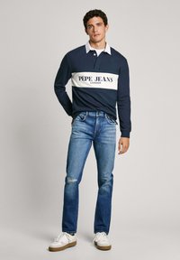 Tamsiai mėlynas ilgomis rankovėmis polo marškinėliai su balta juostele ir "PEPE JEANS LONDON" logotipu; derinami su šviesiai mėlynomis suplėšytomis džinsais ir baltais sportbačiais.
