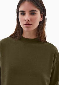 Oliwkowa bluza z wysokim kołnierzem, miękka w dotyku i luźna w kroju. Posiada opadające ramiona oraz gładką, nieozdobioną powierzchnię.