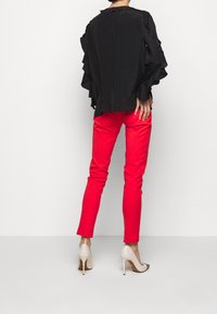 Blouse noire à volants avec un col rond, associée à un pantalon rouge ajusté et des escarpins beiges, mettant en valeur une silhouette élégante.