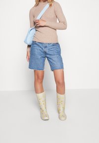 Beige gerippter Pullover, blaue Umhängetasche, blaue Denim-Shorts und beige Gummistiefel mit gelben und pastellfarbigen Punkten. Einfaches Design und lässige Passform.