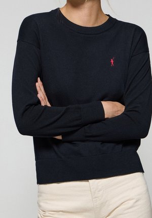 Personne portant un pull à manches longues noir avec un petit logo rouge brodé sur la poitrine, les bras croisés, associée à un pantalon beige clair.