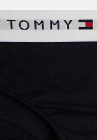 Un slip en tissu noir avec une ceinture blanche sur laquelle est brodé "TOMMY" en noir, accompagné d'un logo carré rouge et bleu.