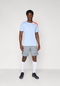 T-shirt de sport Nike bleu clair avec un accent orange sur l'épaule, associé à un short gris avec un logo orange. Chaussures de football noires et chaussettes blanches.