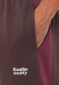 Bordeaux a hnědé tepláky s bočními kapsami, s bílým logem "Kaotiko Society". Měkká tkanina a sportovní design.