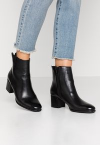 Botas de tobillo de cuero negro con tacón cuadrado, punta afilada y cremallera lateral. Textura lisa, con un diseño de corte bajo en el tobillo.