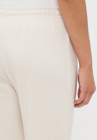 Pantaloni sportivi beige chiaro realizzati in un tessuto morbido, dotati di una vita elastica e di una texture liscia, con una vista ravvicinata della parte laterale e posteriore.