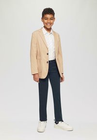 Licht beige blazer over een wit shirt met kleine blauwe patronen, gecombineerd met donker marineblauwe broeken en witte sneakers.