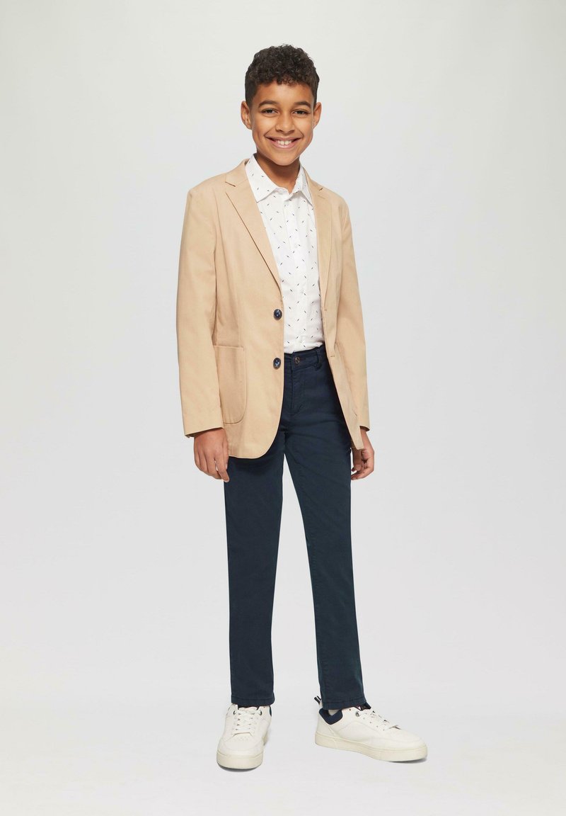 Licht beige blazer over een wit shirt met kleine blauwe patronen, gecombineerd met donker marineblauwe broeken en witte sneakers.