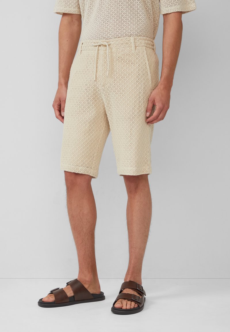 Shorts en maille beige avec une taille à cordon et une coupe décontractée. Associés à des sandales marron foncé avec des doubles brides et une semelle plate.