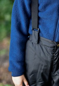 Blauer Fleecepullover mit strukturierter Stoffqualität, kombiniert mit schwarzen Nylonlatzhosen mit verstellbaren schwarzen Trägern und einem dekorativen goldenen Knopf.