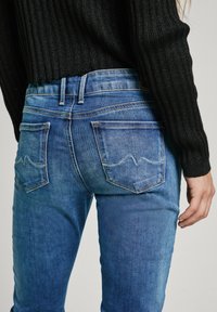 Jeans de mezclilla azul con una cintura de altura media, bolsillos traseros cosidos con un patrón de ola y un ajuste regular. Tejido texturizado con ligeras decoloraciones.