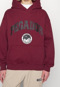 Osoba na sobě má vínovou mikinu s nápisem "PEGADOR" a emblémem hory, nad béžovými kalhotami je nápis "No love lost No love found".