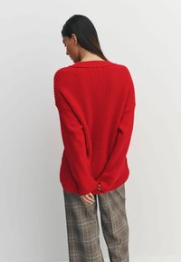 Red knitted sweater with a loose fit and ribbed details, paired with wide plaid trousers in neutral tones. Sweater has a dropped shoulder design.