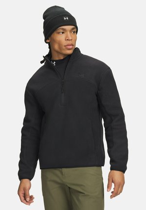 Man draagt een zwarte fleece trui met halve rits en een zwarte muts, kijkt opzij, gekleed in olijfgroene broek.