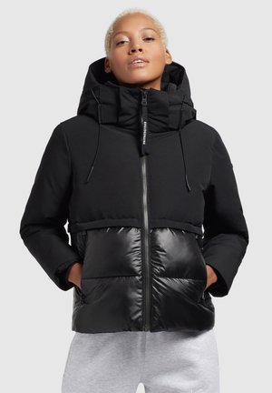 Zwarte jas met een matte bovenkant en glanzende onderkant. Voorzien van een hoge kraag, ritsluiting en twee vo pockets. Vlotte textuur over het hele oppervlak.