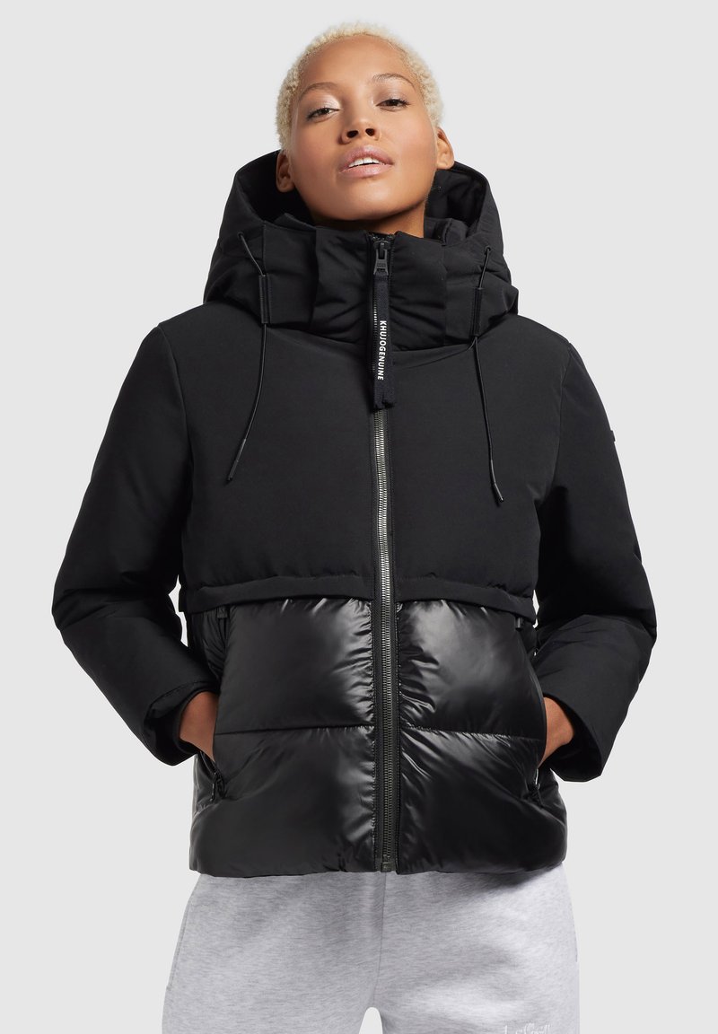 Zwarte jas met een matte bovenkant en glanzende onderkant. Voorzien van een hoge kraag, ritsluiting en twee vo pockets. Vlotte textuur over het hele oppervlak.