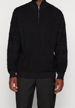 Sort kabelstrikket pullover-trøje med kvartlynlås i krave båret af person med mørke bukser, hænder afslappet langs siderne mod ensfarvet baggrund.